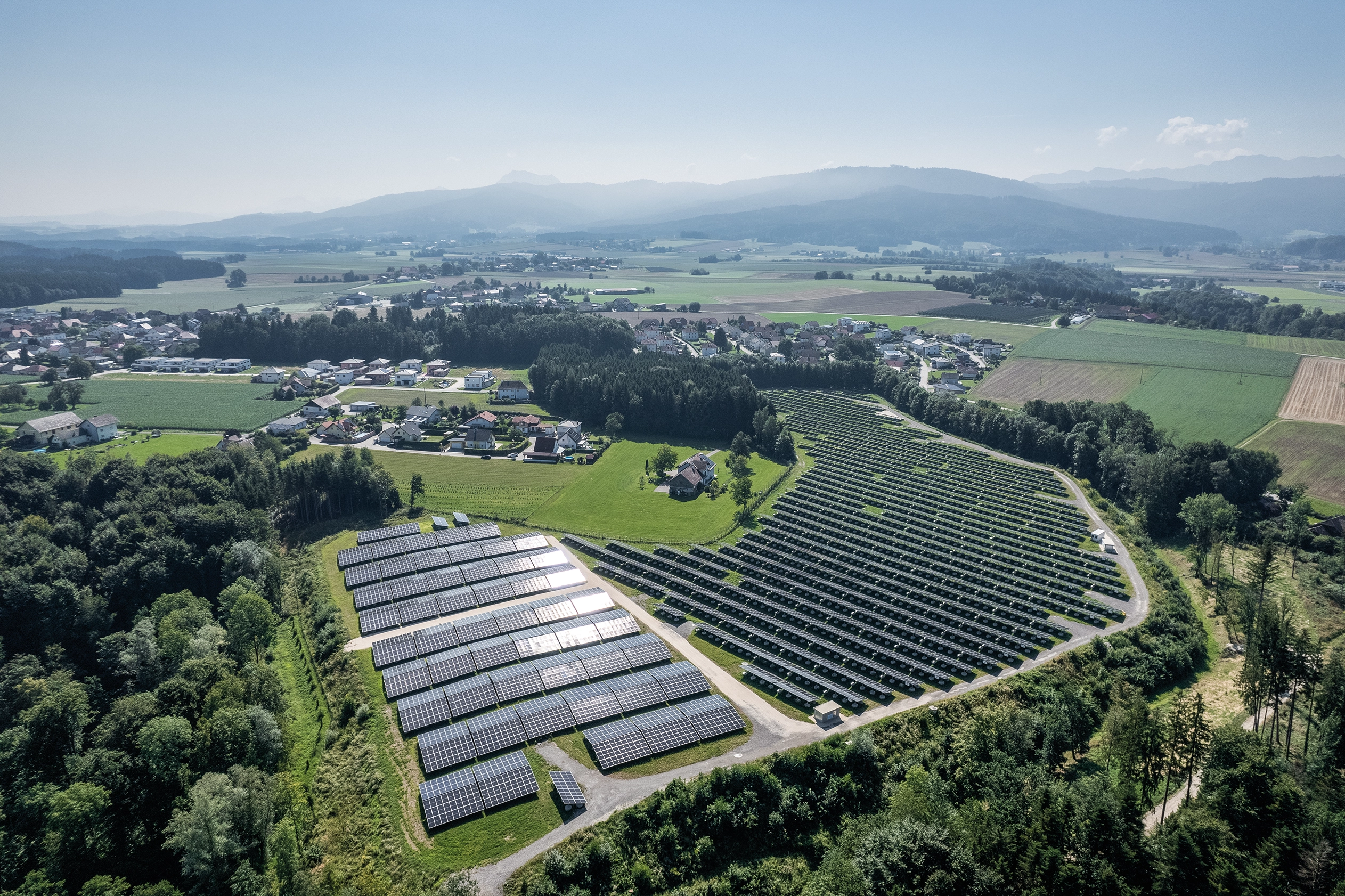 Ein Blick von oben auf eine großflächige Photovotaikanlage. Der Solarpark ist von sattem grün umgeben. Im Hintergrund sieht man kleinere malerische Ortschaften.