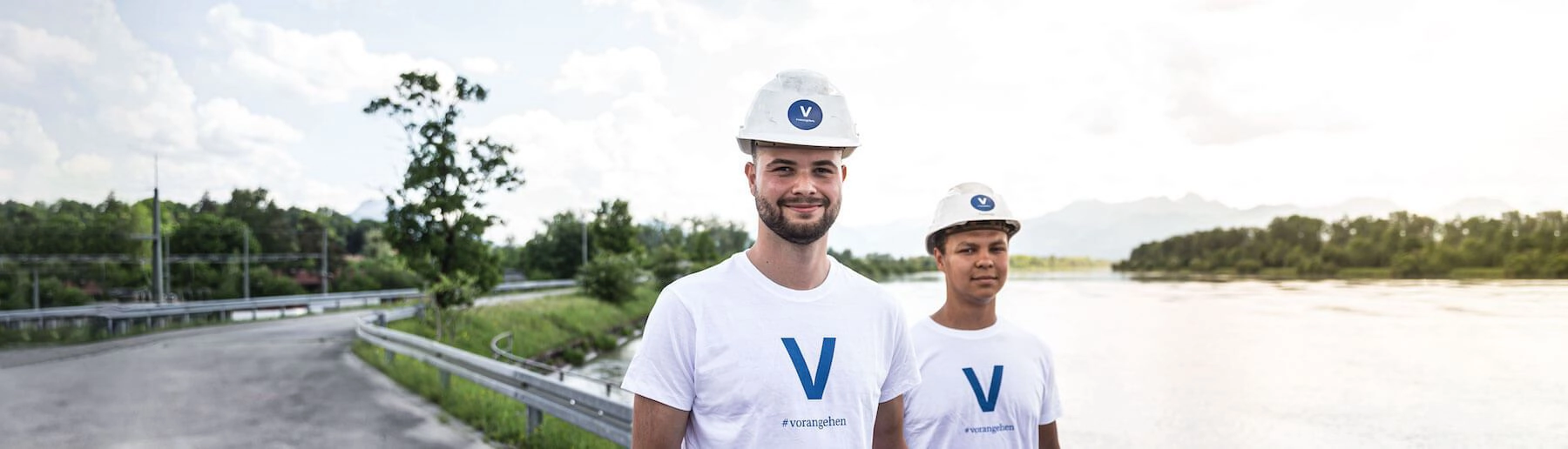 Zwei junge VERBUND-Mitarbeiter in weißen T-Shirts mit blauem V und Helmen lächeln in die Kamera.